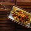 Overhead shot of seasoned french fries topped with chilli sauce and herbs in a metal basket on wooden background.