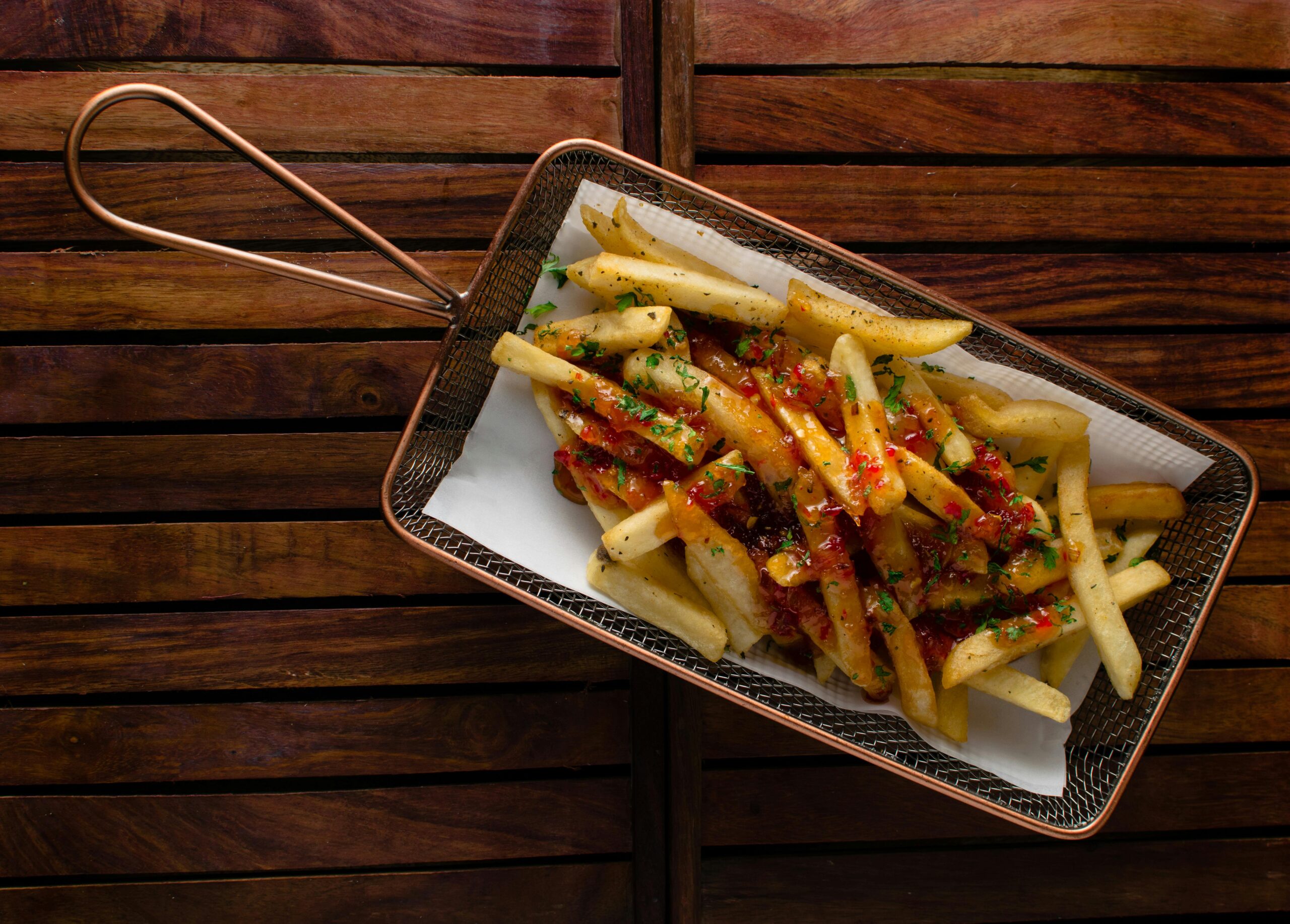 Overhead shot of seasoned french fries topped with chilli sauce and herbs in a metal basket on wooden background.