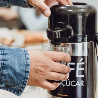 Person pouring coffee from a stainless steel dispenser into a glass.